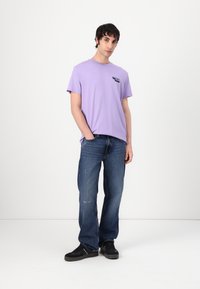 Levandų spalvos medvilninė t-shirt su apvaliu kaklu ir juodu atspauduotu logo, derinama su mėlynomis džinsinėmis kelnėmis su nusidėvėjimu ir juodais sportbotais.