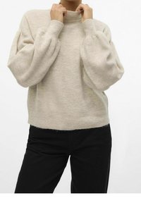 Personne portant un pull côtelé beige à manches bouffantes et un pantalon noir, ajustant le col montant avec les deux mains.
