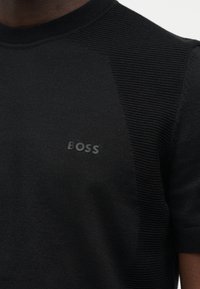 Κοντινή λήψη μιας μαύρης υφασμάτινης κοντομάνικης μπλούζας με το λογότυπο "BOSS" στο στήθος, που φοράει ένα άτομο με σκούρο δέρμα.