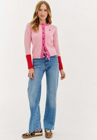 Różowy kardigan z dzianiny z czerwonymi akcentami i guzikami, w połączeniu z niebieskimi dzwonami i butami w panterkę. Posiada wiązanie w talii.