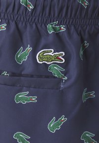 Marineblå shorts med elastisk linning, dekorert med et gjentatt grønt krokodillemønster og et brodert krokodillemerke på lommen.