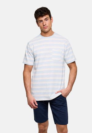 Hombre con camiseta de rayas blancas y azul claro con bolsillo en el pecho y pantalones cortos azul marino, de pie con una mano en el bolsillo.