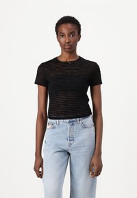 Ung kvinne i en svart gjennomsiktig kortermet topp med tekstmønster og lys blå høytlivte jeans, stående mot en hvit bakgrunn.