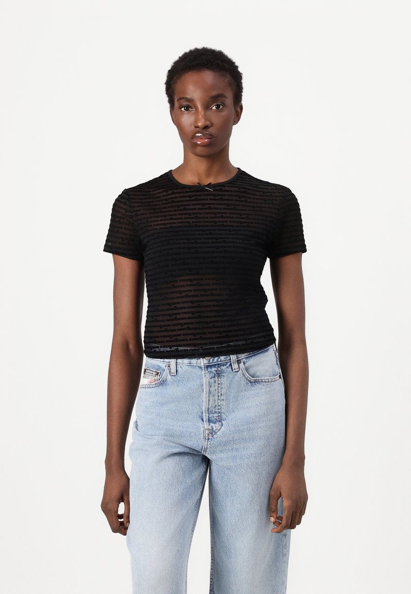 Ung kvinne i en svart gjennomsiktig kortermet topp med tekstmønster og lys blå høytlivte jeans, stående mot en hvit bakgrunn.