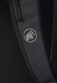 Svart stoff med et teksturert rutenettmønster; har et sølvfarget elefantlogo. Inkluderer en solid stropp for festing.