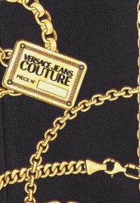 Tissu noir avec un imprimé de chaînes dorées et une étiquette rectangulaire portant la mention "Versace Jeans Couture" en lettres grasses.