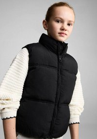 Vest puffer negru cu guler înalt și fermoar frontal. Are un design matlasat și o textură moale, purtat peste un pulover tricotat alb.