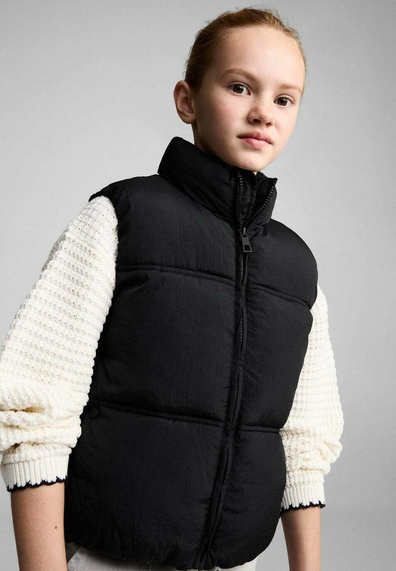 Vest puffer negru cu guler înalt și fermoar frontal. Are un design matlasat și o textură moale, purtat peste un pulover tricotat alb.