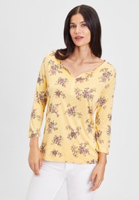 Gelbes Langarmshirt mit V-Ausschnitt, ausgestattet mit floralen Mustern in Lila und Pink. Weiches Material mit einem Knopfdetail entlang des Ausschnitts.