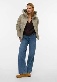 Veste beige rembourrée avec une grande capuche, associée à un pull en tricot foncé et un jean bleu à jambes larges. Chaussures marron avec semelles crantées visibles.