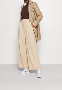 Pantalones de pierna ancha beige con una cintura alta, combinados con un suéter marrón y un abrigo marrón claro, completados con zapatillas blancas.