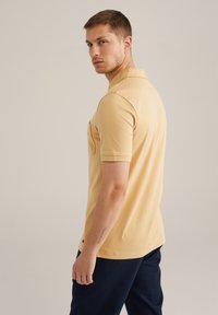 Tricou polo din bumbac galben deschis, cu mâneci scurte, guler și un buzunar pe piept, având o croială regulară și accente pe marginile laterale.