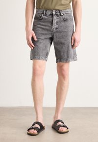 Shorts en denim noir à coupe droite, avec un ourlet effiloché, cinq poches et une fermeture à bouton, associés à des sandales noires à enfiler sur un fond neutre.