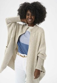 Beżowy sweter z dzianiny o prążkowanym wzorze, otwarty z przodu, z bocznymi kieszeniami. Nosi się go na niebieską i białą koszulę w paski oraz białe spodnie.