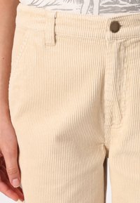 Beige corduroy pants feature vertical ribbing, a front button, and a simple waistband. The fabric has a soft, textured finish.