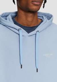 Lichtblauwe hoodie met verstelbare trekkoorden, metalen uiteinden en bedrukte "STREET ART" logo. Zachte stoftextuur, relaxte pasvorm, over een donkere shirt gelaagd.