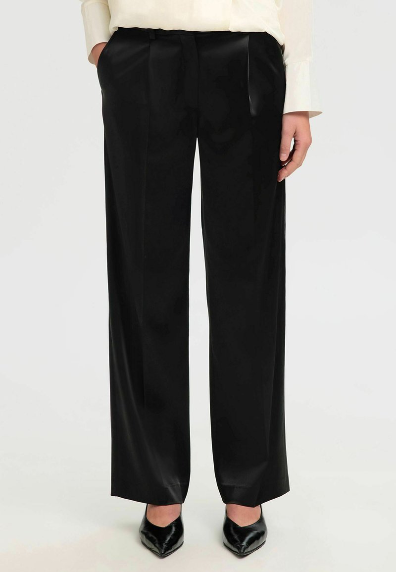 Pantalons larges en satin noir avec une texture lisse, des plis sur le devant et une taille haute, associés à des chaussures noires à bout pointu.