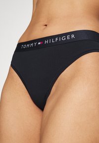 Crna pamučna tanga gaćice s elastičnom trakom s logom na kojoj piše "TOMMY HILFIGER" u bijeloj i ružičastoj boji. Glatka tekstura s minimalnim dizajnom.