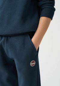 Tamsiai mėlynas džemperis ir kelnes iš minkštos medžiagos. Kelnės turi šoninį kišenę ir apvalų logotipo pleistrą. Rankogaliai yra su gūsiu, kad gerai priglustų.