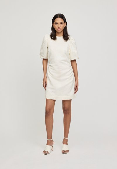 Crème-kleurige jurk met gepofte korte mouwen, gerimpeld zijdetail en een rechte silhouet, gecombineerd met witte hoge hakken sandalen.