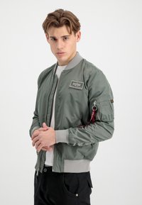 Olivgrön bomberjacka i glänsande nylon, med grått ribbat manschetter och nederkant, zip-stängning och en broderad logotyp på bröstet. Justerbara fickdetaljer.
