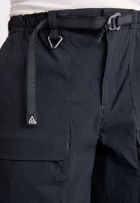 Black cargo pants made from lightweight fabric, featuring a woven belt, zip and flap pockets, and a triangular logo patch.