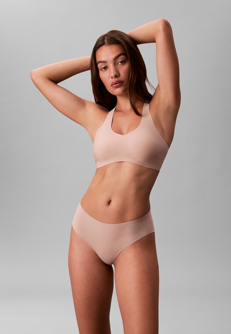 Nude sømløs bralette med rund halsudskæring og brede stropper, kombineret med matchende lavtaljede trusser. Blødt, elastisk stof med minimal detaljering.