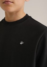 Svart sweatshirt med rund hals, prydd med en liten vit fågel-emblem på bröstet. Tillverkad av mjukt bomullsmaterial.