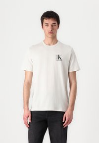 Bílý krátký rukáv tričko z měkké bavlny, s malým šedým logem "CK" na hrudi. Model spárovaný s tmavými džíny.