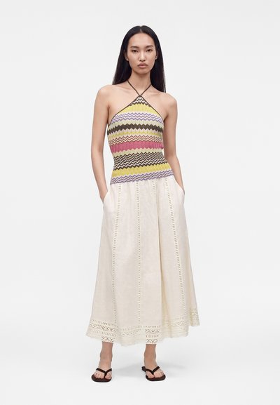 Γυναίκα που φοράει ένα αμάνικο halter top με ζιγκ-ζαγκ μοτίβο και κίτρινες, ροζ και μωβ ρίγες, συνδυασμένο με μια κρεμ μακριά φούστα και μαύρα σανδάλια.