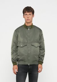 Giacca bomber verde oliva con chiusura a zip, collo e polsini a costine, due tasche frontali con pattina e una finitura liscia e lucida.