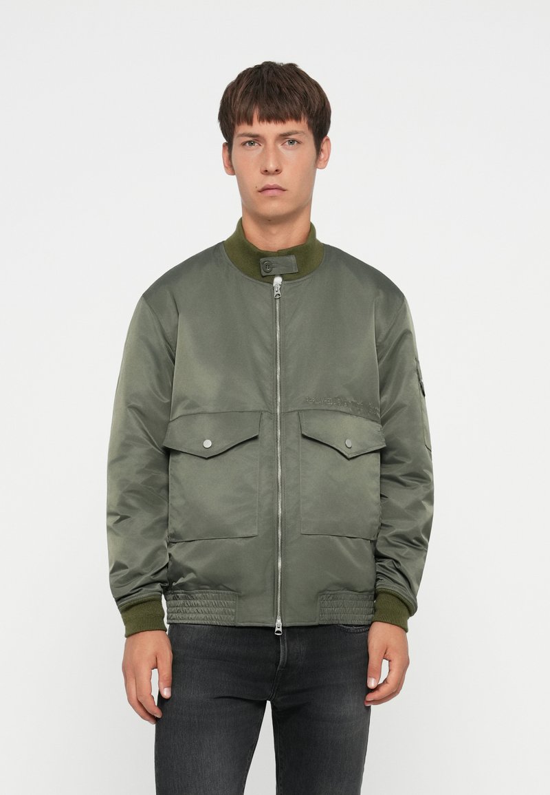 Giacca bomber verde oliva con chiusura a zip, collo e polsini a costine, due tasche frontali con pattina e una finitura liscia e lucida.