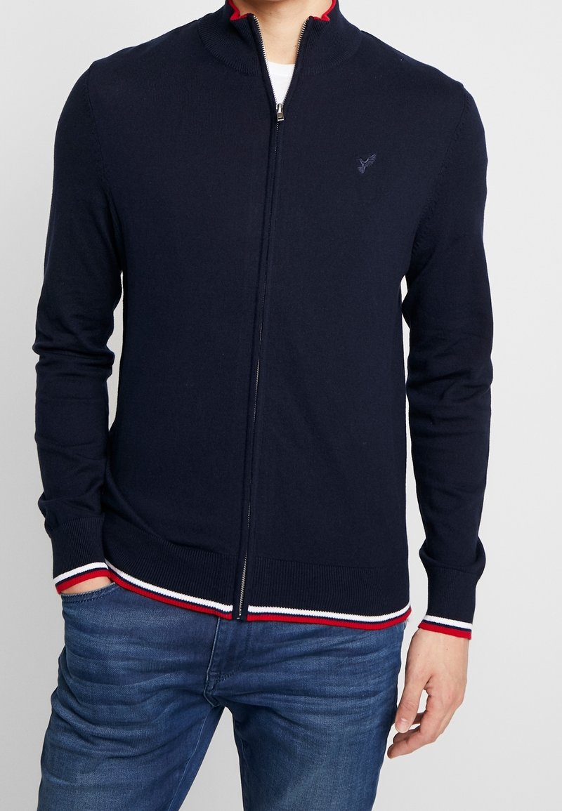 Man som bär en marinblå zip-up tröja med röd, vit och blå randig design på ärmsluten och nederkanten, ihop med blå jeans, med avslappnade händer.