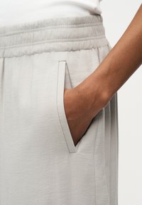 Main dans la poche latérale d'un pantalon beige clair à taille élastique, porté avec un haut blanc, montrant la texture du tissu et les détails des coutures.