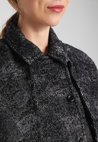 Černý a šedý kabát z vlněného materiálu s vzorem houndstooth, vysokým límcem, zapínáním na knoflíky a texturovaným povrchem.