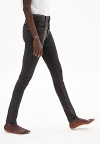 Personne marchant pieds nus portant un jean skinny gris foncé et une chemise blanche, montrée de la moitié du torse jusqu'aux pieds sur un fond blanc.