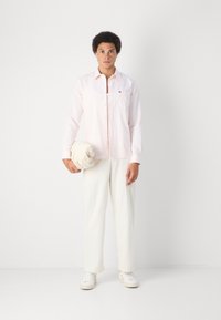 Camicia a maniche lunghe rosa chiaro con taschino e logo; abbinata a pantaloni crema e tiene un tessuto beige arrotolato, sneakers bianche.