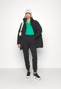 Giubbotto piumino nero, polo a coste verde, pantaloni jogger grigi e sneaker nere con suola bianca; modello che indossa un cappellino bianco.