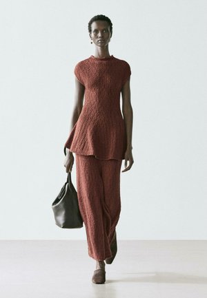 Kobieta idąca do przodu, ubrana w teksturowaną tunikę w kolorze rdzy i szerokie spodnie, niosąca czarną torebkę, na tle jasnego, jednolitego tła.