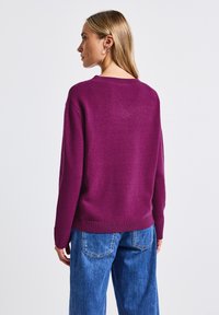 Pull violet avec un col rond et des poignets côtelés. Porté avec un jean bleu à jambe large, mettant en valeur une texture tricotée et une coupe décontractée.