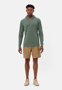Felpa verde a maniche lunghe con tasca a marsupio, abbinata a pantaloni corti beige. Le calzature sono grigie chiare con dettagli rossi. Tessuto dalla consistenza liscia.