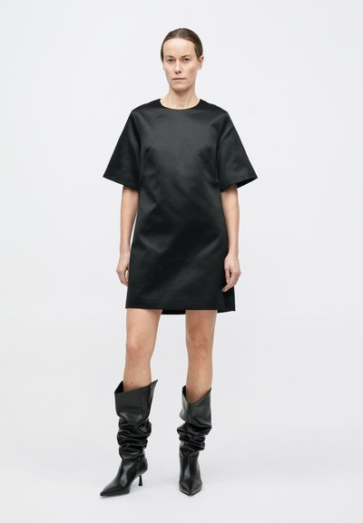 Mini-robe en satin noir avec manches courtes et encolure ronde. Associée à des bottes en cuir noir bouffantes montant jusqu'aux genoux. Texture lisse.