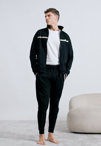 Czarny sportowy dres z kurtką na suwak z białym i złotym paskiem, w zestawie ze zwężanymi spodniami, na jasnym tle.