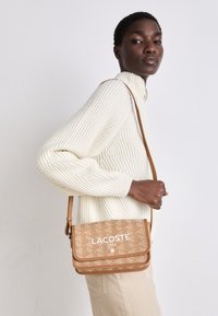 Borsa a tracolla marrone con un motivo geometrico, caratterizzata dal testo del logo bianco "LACOSTE 1933". Materiale liscio, tracolla regolabile e chiusura a pattina.