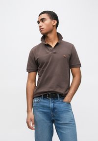 Polo castanha com colarinho dobrado e um pequeno logótipo, combinada com jeans azul claro. O tecido aparenta ser liso com um corte descontraído.