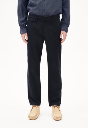 Uomo che indossa pantaloni dritti blu scuro, camicia blu scuro con bottoni e mocassini beige, in piedi su sfondo bianco.
