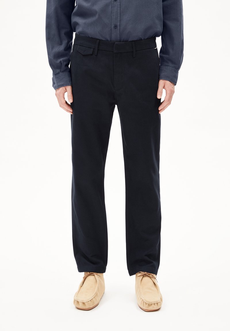 Homme portant un pantalon droit bleu marine foncé, une chemise bleu marine à boutons et des mocassins beige, debout devant un fond blanc.
