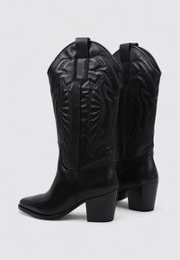 Bottes de cowboy en cuir noir avec un bout pointu et un talon de hauteur moyenne. Elles présentent une couture décorative sur l'empeigne et un tirant.