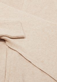 Beige geribde gebreide trui met opgestroopte mouwen op een bijpassende geribde stofachtergrond.