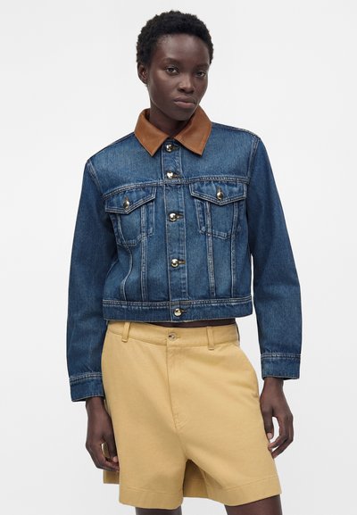 Γυναίκα που φορά μπλε τζιν μπουφάν με καφέ γιακά και μπεζ σορτς, στέκεται μπροστά σε απλό λευκό φόντο.